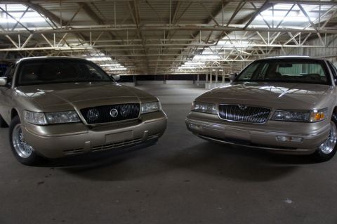 2000 Mercury Grand Marquis - Two Mercury Grand Marquis sedans are parked side-by-side in a large indoor space with exposed beams overhead.