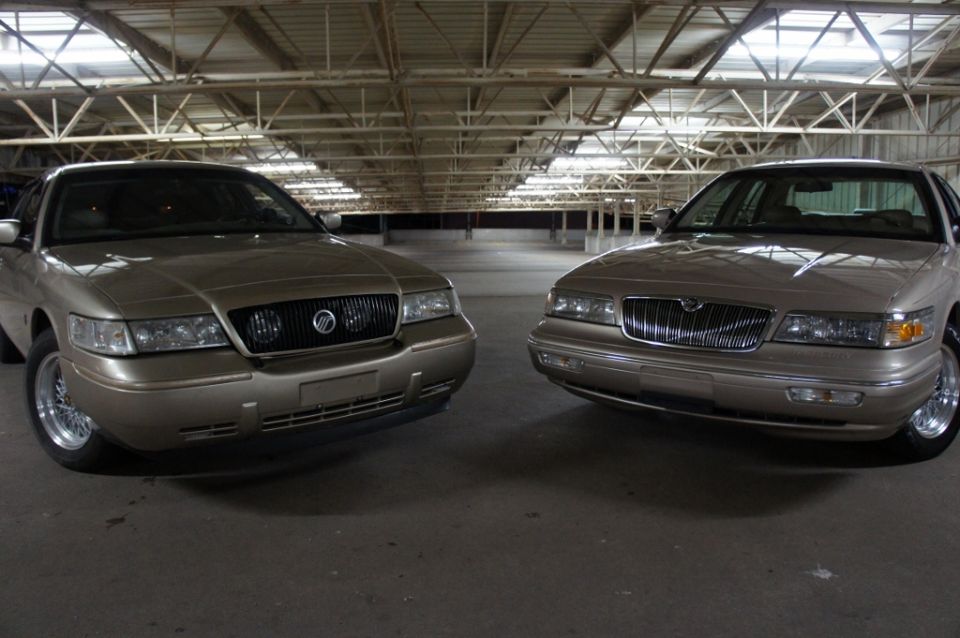 Two tan Mercury Grand Marquis sedans parked inside a large building. The one on the left has a black grille, the one on the right has a chrome grille