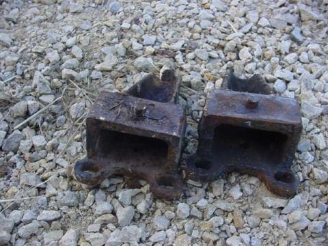 1992 Ford Bronco - Two old, rusted metal brackets sit on a bed of gravel and dry grass.