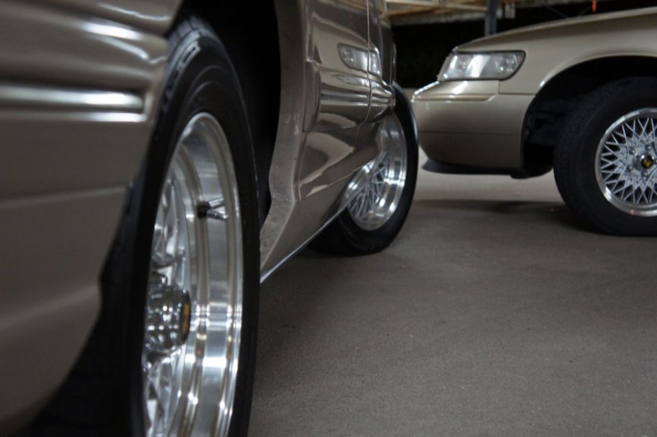 Two cars parked, close-up on the front wheel and side panel of the car on the left, and the front wheel and headlight of the car on the right. The car on the right appears to be a 2000 Mercury Grand Marquis