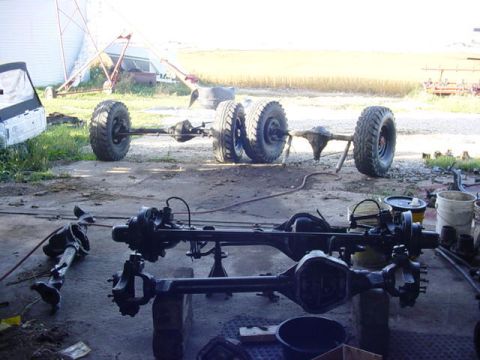 1992 Ford Bronco - Multiple vehicle axles and large tires are laid out on a dirt surface, with a partial view of a white truck cab in the background.