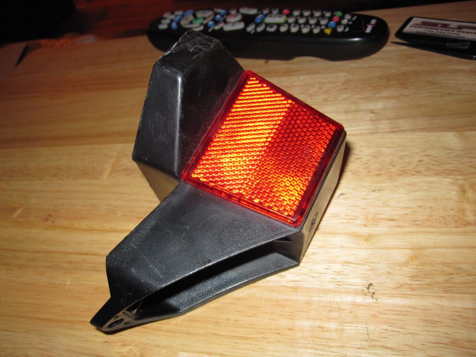 Motorcycle tail light assembly with red reflector, sitting on a wooden surface next to a remote control