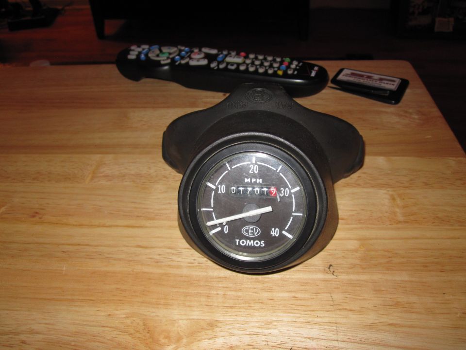 Motorcycle speedometer with 'TOMOS' logo, reads 01707.9 miles. Remote control and cassette tape nearby