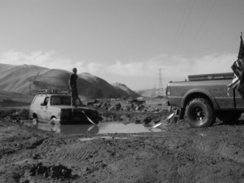 1990 Ford Bronco - A 1990 Ford Bronco is stuck in the mud, with a person standing on its hood, while a pickup truck is parked nearby.