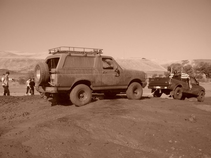 Muddy Ford Bronco with roof rack and spare tire on back, towing a pickup truck with American flags