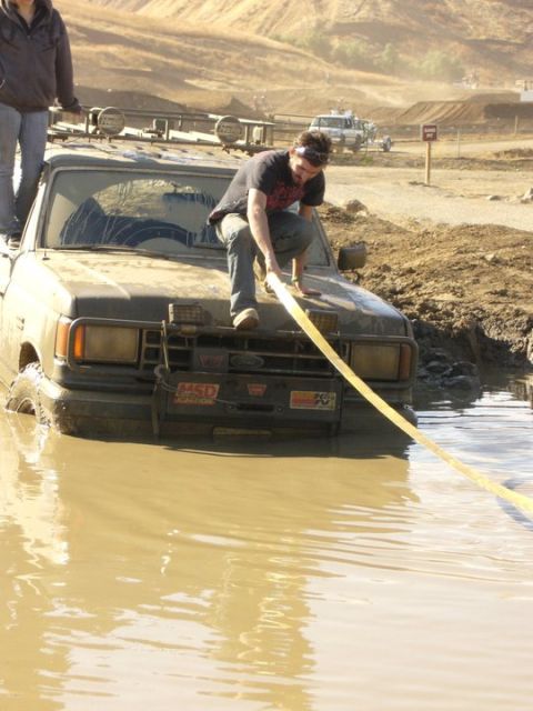 1990 Ford Bronco - A muddy Ford Bronco is stuck in water while two people attempt to pull it out with a strap.