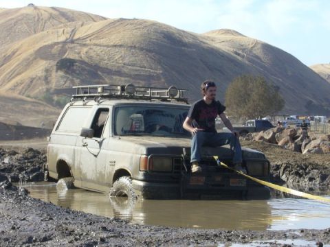 1990 Ford Bronco - A 1990 Ford Bronco is stuck in deep mud, with a person sitting on its hood and a tow strap attached to the front.
