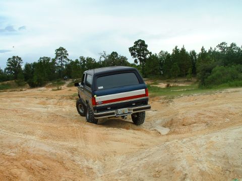 1995 Ford Bronco - A dark blue SUV navigates a challenging dirt terrain with trees in the background.