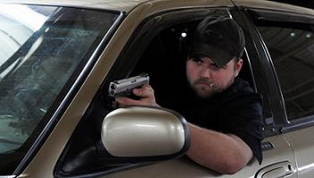 2000 Mercury Grand Marquis - A man holds a handgun out the window of a tan car, looking towards the camera.