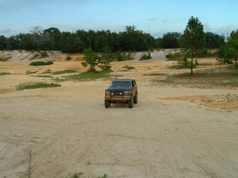 1995 Ford Bronco - A dark-colored Ford Bronco sits in a sandy clearing, surrounded by trees and uneven terrain under a cloudy sky.