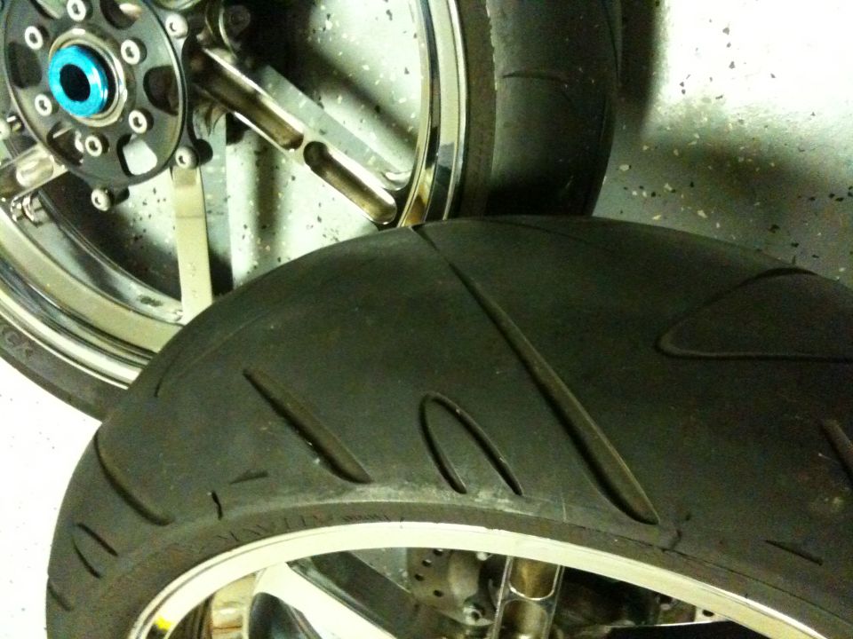Close-up of two motorcycle tires, one in front of the other. The rear tire is larger and shows tread detail. The front wheel has a blue center cap