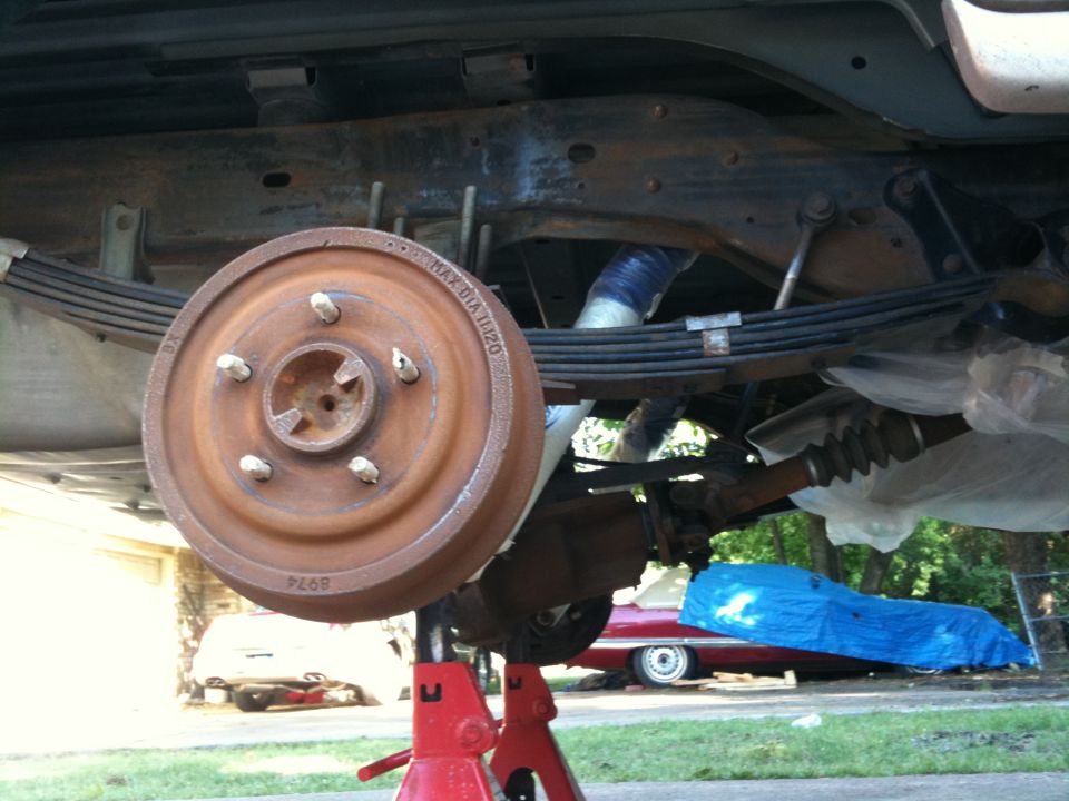 Underside of a vehicle on jack stands, showing the rear axle, leaf springs, and brake drum. Another car is visible in the background