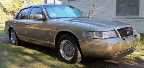 2000 Mercury Grand Marquis - A gold 2000 Mercury Grand Marquis is parked on a grassy area next to a building.