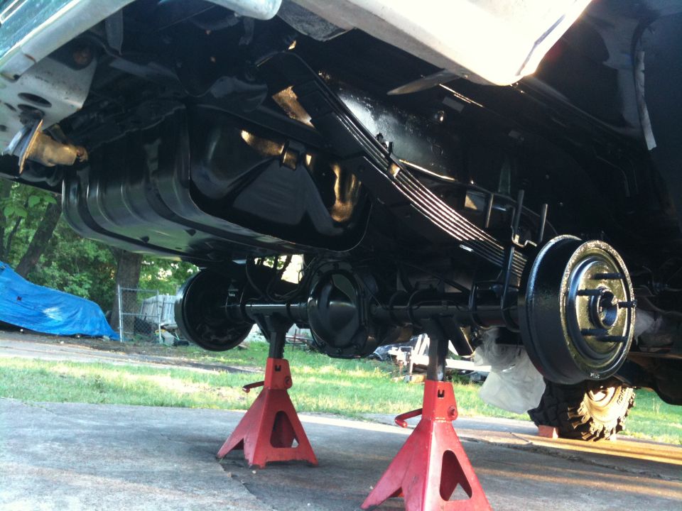 Undercarriage of a vehicle, showing the rear axle, leaf springs, and fuel tank, supported by red jack stands