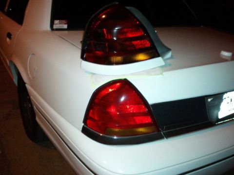 1998 Ford Crown Victoria - Night shot of the rear corner of a white sedan with a white lip spoiler taped to the trunk lid, masking tape still along its edge. A spare…