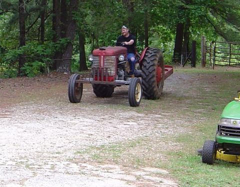1986 Ford Bronco - A man drives a vintage red tractor on a dirt road, with a modern lawnmower visible on the side.