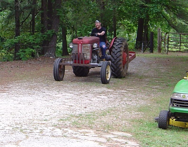 Man driving a red tractor on a gravel path, with trees in the background and a lawnmower to the right
