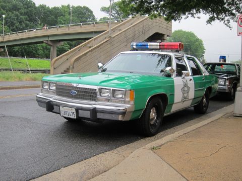 1991 Ford LTD Crown Victoria - green and white Ford LTD Crown Victoria sheriff sedan with a roof light bar is parked along a road beside a pedestrian overpass.