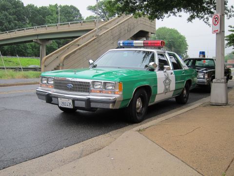 1991 Ford LTD Crown Victoria - green and white Ford LTD Crown Victoria sheriff cruiser with a red and blue light bar sits parked at a curb near a highway overpass.