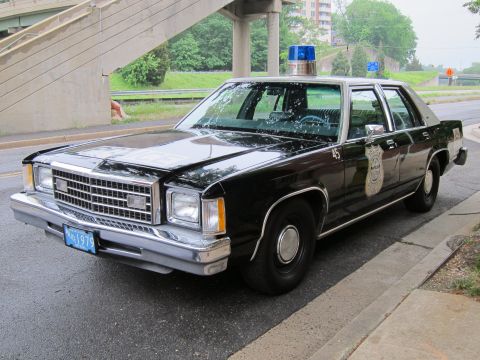 1991 Ford LTD Crown Victoria - black Ford LTD Crown Victoria police sedan with a single blue roof beacon and gold door emblem is parked along a road near a highway overpa…