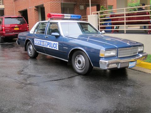 1991 Ford LTD Crown Victoria - blue Chevrolet Caprice marked 'North Haven Police' with a roof light bar is parked on wet pavement beside a red brick building.