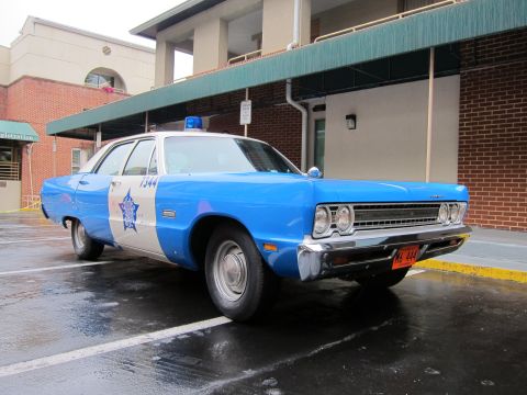 1991 Ford LTD Crown Victoria - blue and white vintage police sedan marked 1344 with a star emblem and roof beacon is parked on wet pavement outside a building.