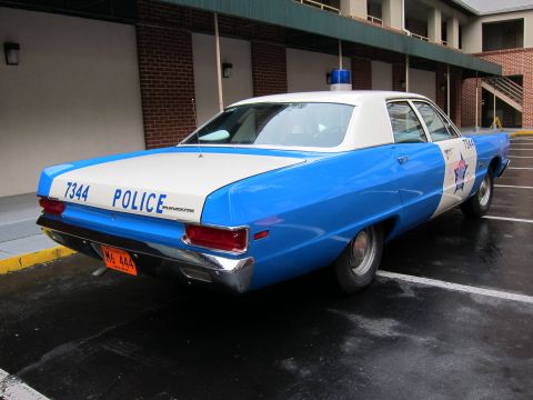 1991 Ford LTD Crown Victoria - Rear three-quarter view of a blue and white vintage police sedan marked '7344 POLICE' with a star emblem and roof beacon, parked on wet pav…