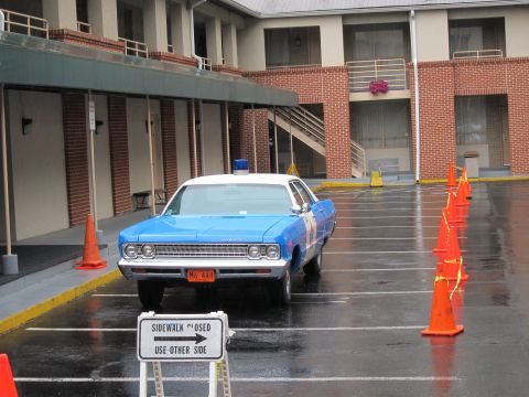 1991 Ford LTD Crown Victoria - blue-and-white classic police sedan with a roof light bar sits in a rain-soaked motel parking lot lined with orange cones.
