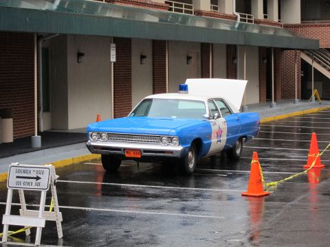 1991 Ford LTD Crown Victoria - blue-and-white vintage police cruiser bearing a star emblem parked on wet pavement beside a sidewalk-closed sign and orange cones.