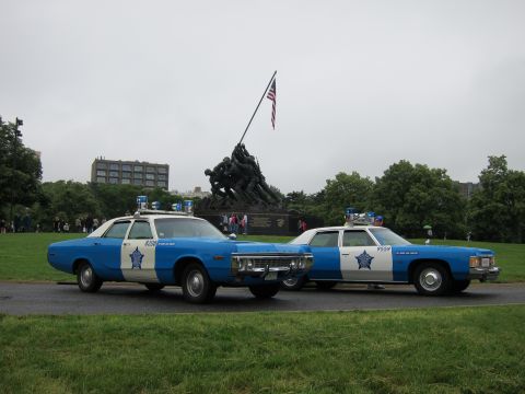 1991 Ford LTD Crown Victoria - Two blue-and-white vintage police sedans with roof lights parked side by side on grass before the Iwo Jima Memorial statue.