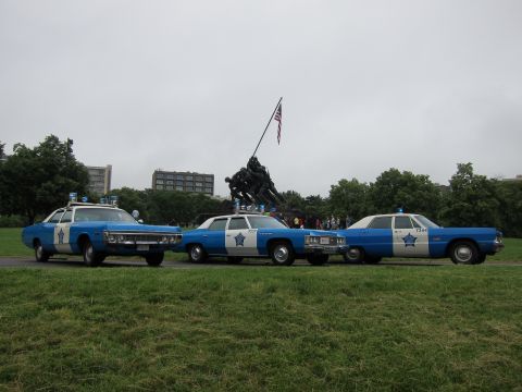 1991 Ford LTD Crown Victoria - Three blue-and-white vintage police sedans parked in a row on grass with the Iwo Jima Memorial statue behind them.