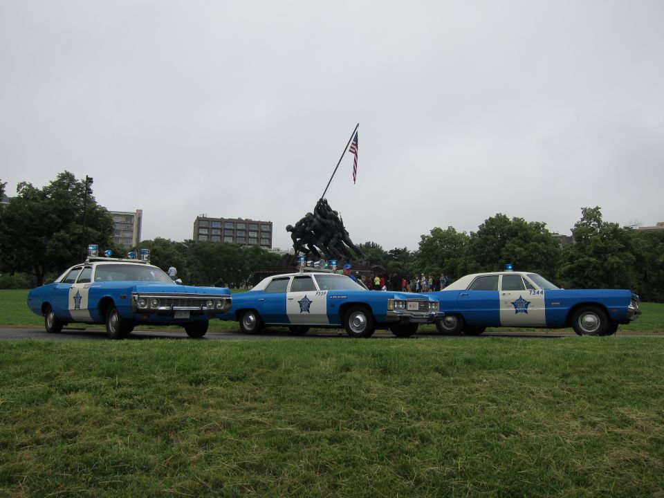 Three blue and white classic police cars lined up before the Iwo Jima Memorial