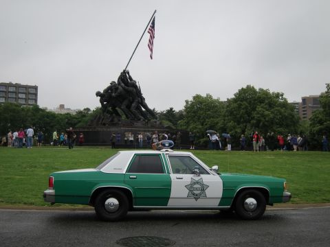 1991 Ford LTD Crown Victoria - green-and-white sheriff sedan with roof lights parked on a road before the Iwo Jima Memorial surrounded by spectators.