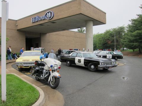 1991 Ford LTD Crown Victoria - police Harley-Davidson motorcycle and several classic black-and-white police cars gathered under a Hilton hotel portico.
