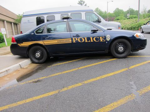 1991 Ford LTD Crown Victoria - Side view of a dark-blue police Impala with a yellow stripe and POLICE markings parked along a curb near a shuttle van.
