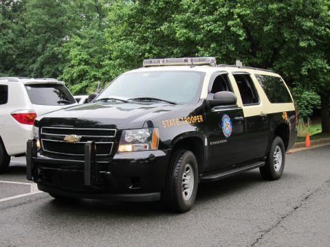 1991 Ford LTD Crown Victoria - black-and-tan Chevrolet Suburban marked State Trooper with a roof light bar and brush guard parked among trees.