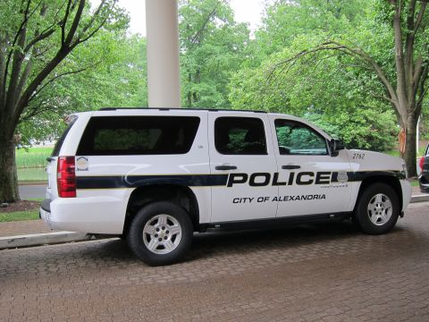 1991 Ford LTD Crown Victoria - white Chevrolet Suburban marked POLICE City of Alexandria with a blue stripe parked on a brick driveway under a canopy.