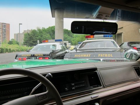 1991 Ford LTD Crown Victoria - View through a car windshield of two black Highway Patrol cruisers with roof lights parked under a building overhang.