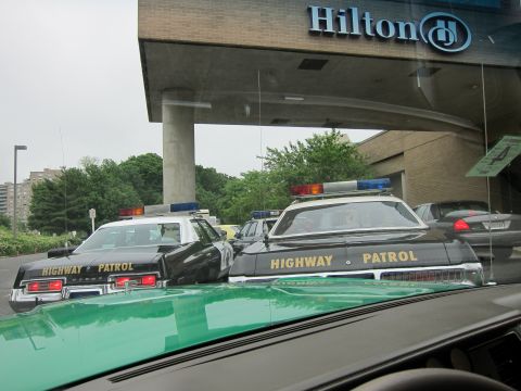 1991 Ford LTD Crown Victoria - Two black Highway Patrol cruisers with light bars parked under the Hilton entrance, seen over the green hood of another car.