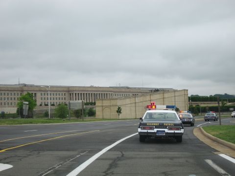 1991 Ford LTD Crown Victoria - black-and-white police sedan marked HIGHWAY PATROL travels a road past the long facade of the Pentagon under a cloudy sky.