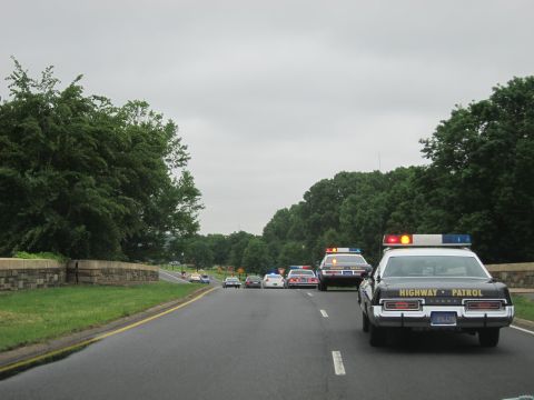 1991 Ford LTD Crown Victoria - black-and-white HIGHWAY PATROL cruiser with roof lights leads a line of cars along a tree-lined two-lane road.