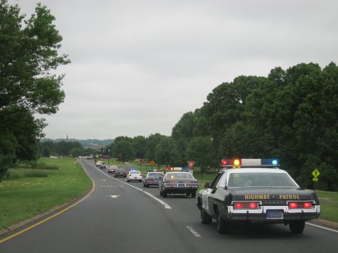 1991 Ford LTD Crown Victoria - black-and-white HIGHWAY PATROL sedan with lit roof bar travels a rural road, a distant spire visible on the horizon.