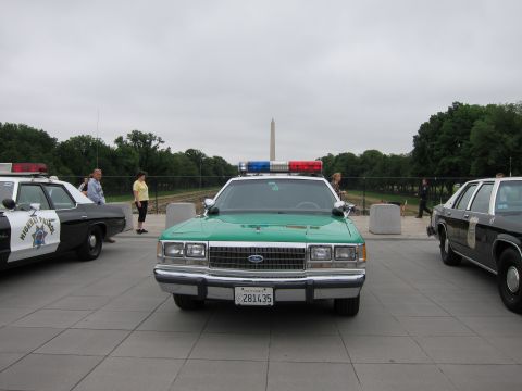 1991 Ford LTD Crown Victoria - green-and-white Ford police sedan with roof lightbar is parked on a plaza, the Washington Monument rising behind it.