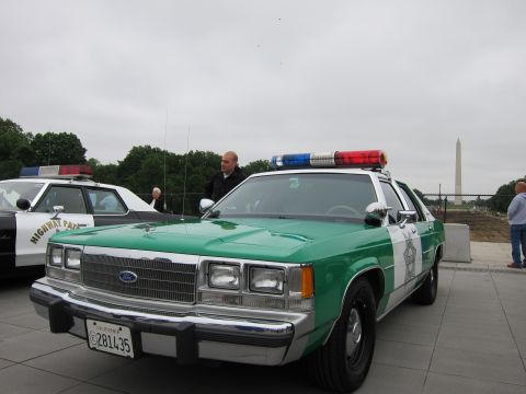 1991 Ford LTD Crown Victoria - man stands beside a green-and-white Ford police sedan with roof lights, the Washington Monument visible behind on a gray day.