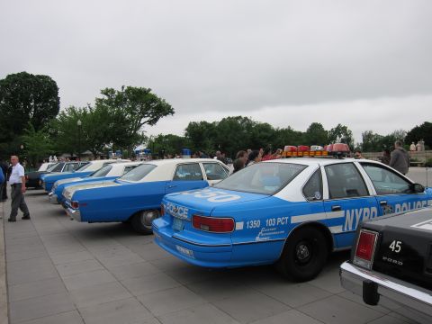 1991 Ford LTD Crown Victoria - row of vintage blue-and-white NYPD police cars is parked on a plaza, a Ford cruiser in the foreground, others lined behind.