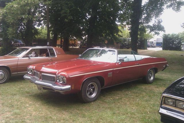 Red convertible with white interior parked next to a brown sedan and a black car. Trees and grass in background