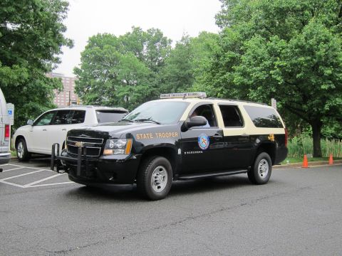 1991 Ford LTD Crown Victoria - black Chevrolet Suburban marked STATE TROOPER with a roof lightbar is parked in a paved lot beside trees.