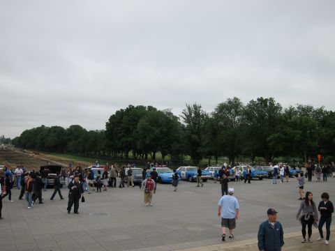 1991 Ford LTD Crown Victoria - Visitors stroll across a large plaza where a row of blue-and-white police cars is parked under an overcast sky.