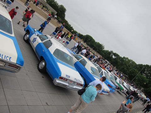 1991 Ford LTD Crown Victoria - diagonal row of vintage blue-and-white POLICE cars is parked on a plaza as a crowd of people gathers behind them.