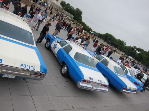 1991 Ford LTD Crown Victoria - Several vintage blue-and-white POLICE cruisers are lined up rear-to-camera on a plaza with onlookers walking past.